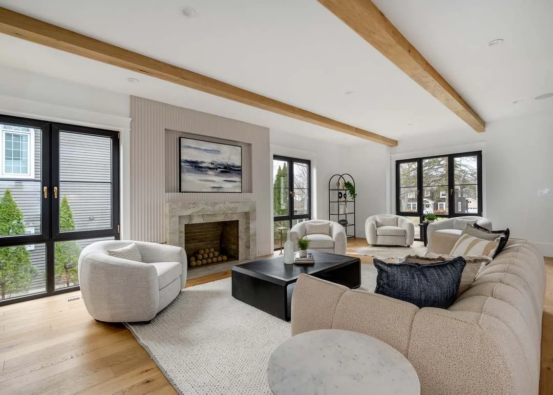 Bright living room with large windows, white sofa, and armchairs, a black coffee table, a fireplace with a painting above, wooden beams on the ceiling, and light wooden flooring.
