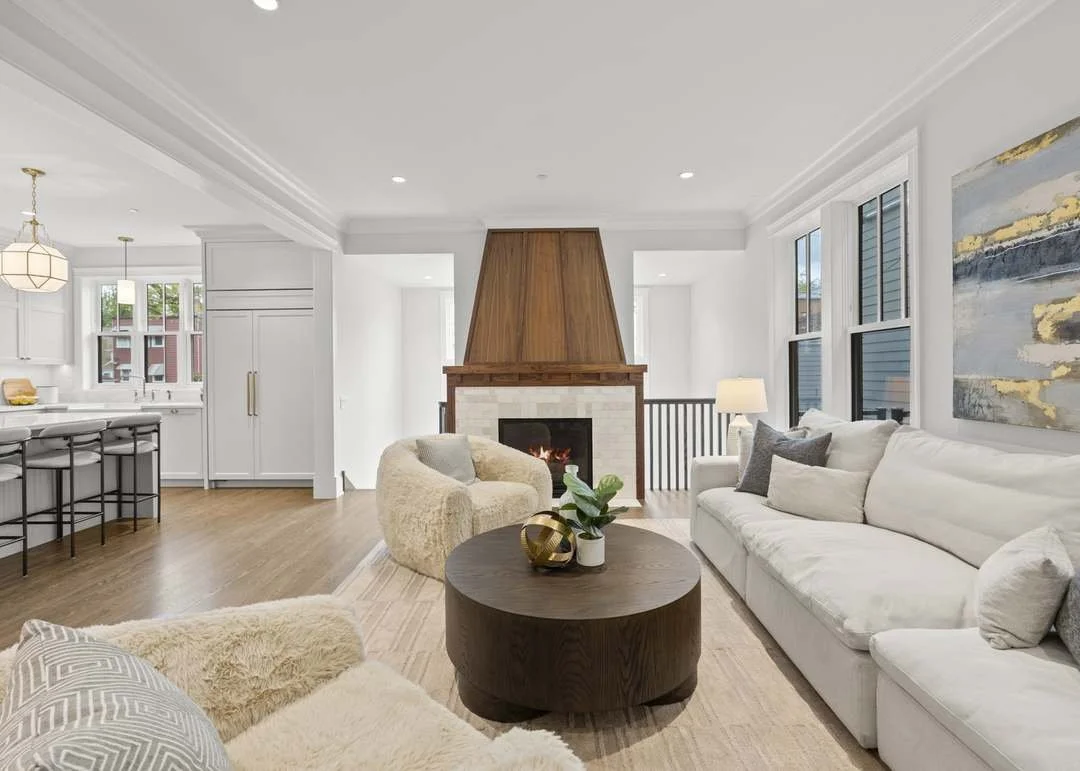Bright living room with white walls, large windows, a white sofa, a beige armchair, a round wooden coffee table with decorative items, a fireplace with a wooden mantel, and a view into the kitchen area.