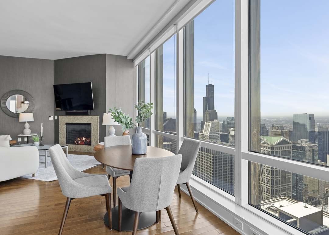 Living room with large windows overlooking a cityscape, includes a round dining table, four chairs, a fireplace, a flat-screen TV, and modern decor.