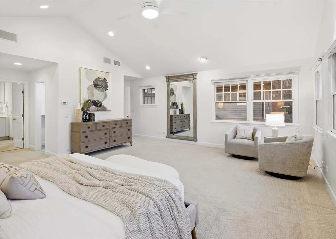 Bright, modern bedroom with white walls, a large bed with neutral-colored bedding, a wooden dresser, a large mirror, and two light gray armchairs near windows with blinds. There are decorative pillows and a lamp on one armchair. The room is illuminated by ceiling lights.