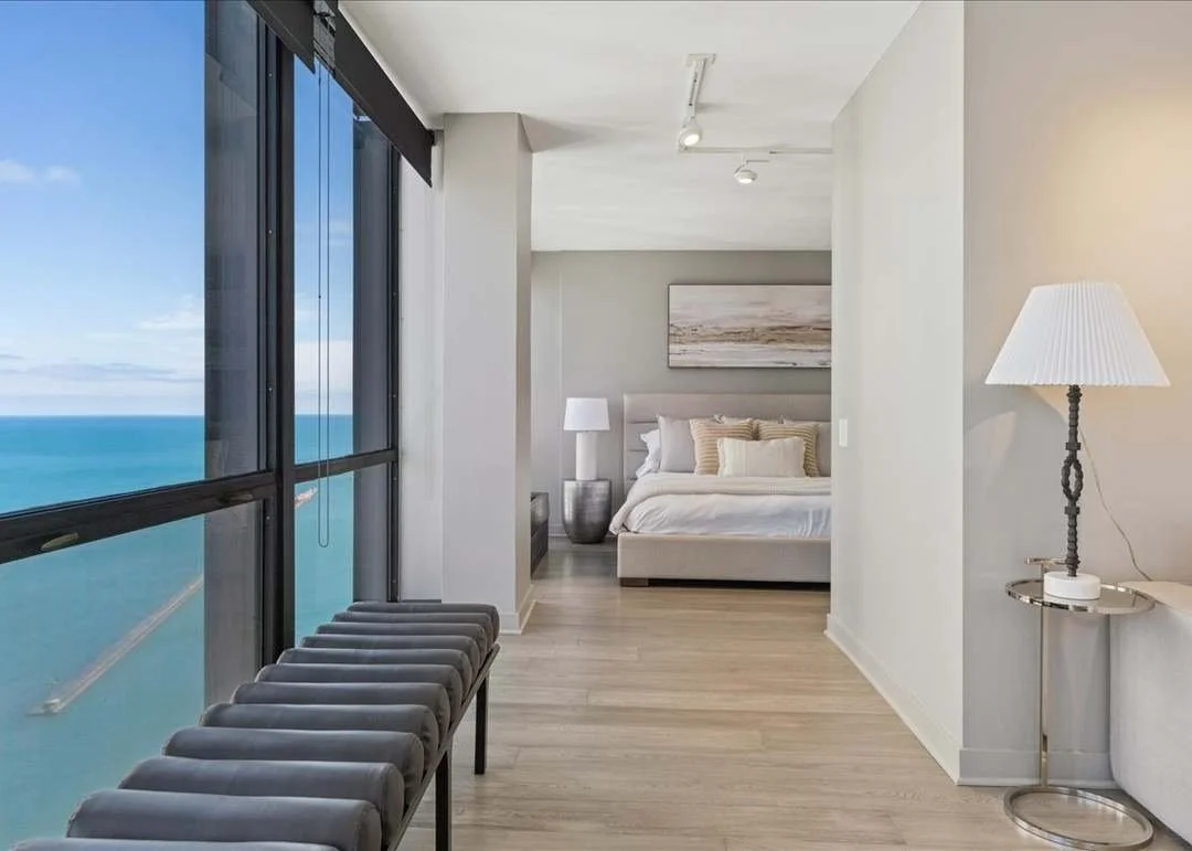 Modern bedroom with large floor-to-ceiling windows showing a view of the ocean, white bedding, a nightstand with a lamp, and minimalistic decor.