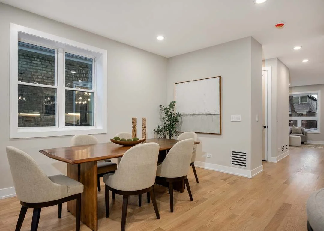 Dining room with a wooden table, six beige upholstered chairs, a decorative bowl, candles, a plant, a large white abstract painting, a window with a brick exterior, and hardwood flooring.