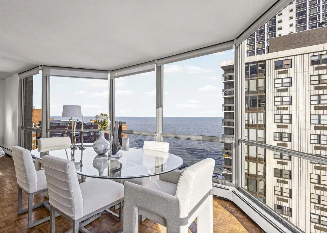 Modern dining room with glass table, six white upholstered chairs, decorative vases, and a view of the city skyline and ocean through large curved windows.