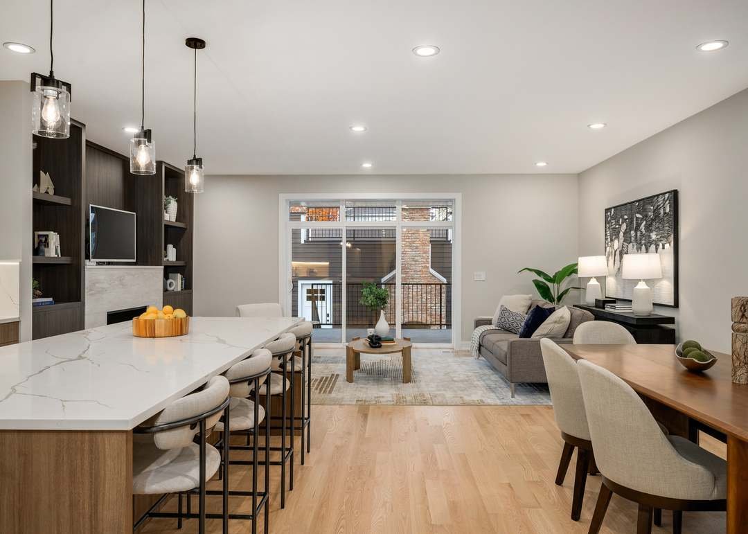 Modern living room with a kitchen island, barstools, a sitting area with a gray sofa, coffee table, sliding glass door, and decorative elements like lamps, plants, and artwork.