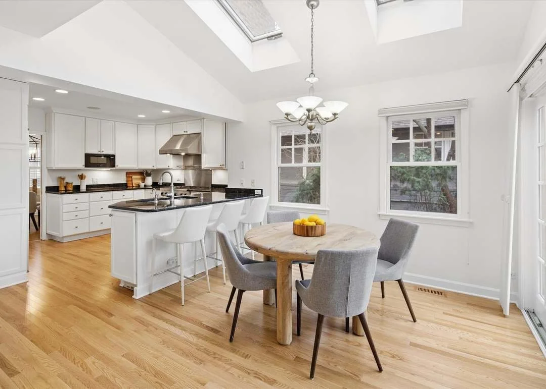 Bright, open kitchen and dining area with white cabinets, black countertops, and wooden floors. Includes a round wooden dining table with four gray chairs and a chandelier. Kitchen has a stainless steel range hood and microwave, with large windows and a skylight letting in natural light.