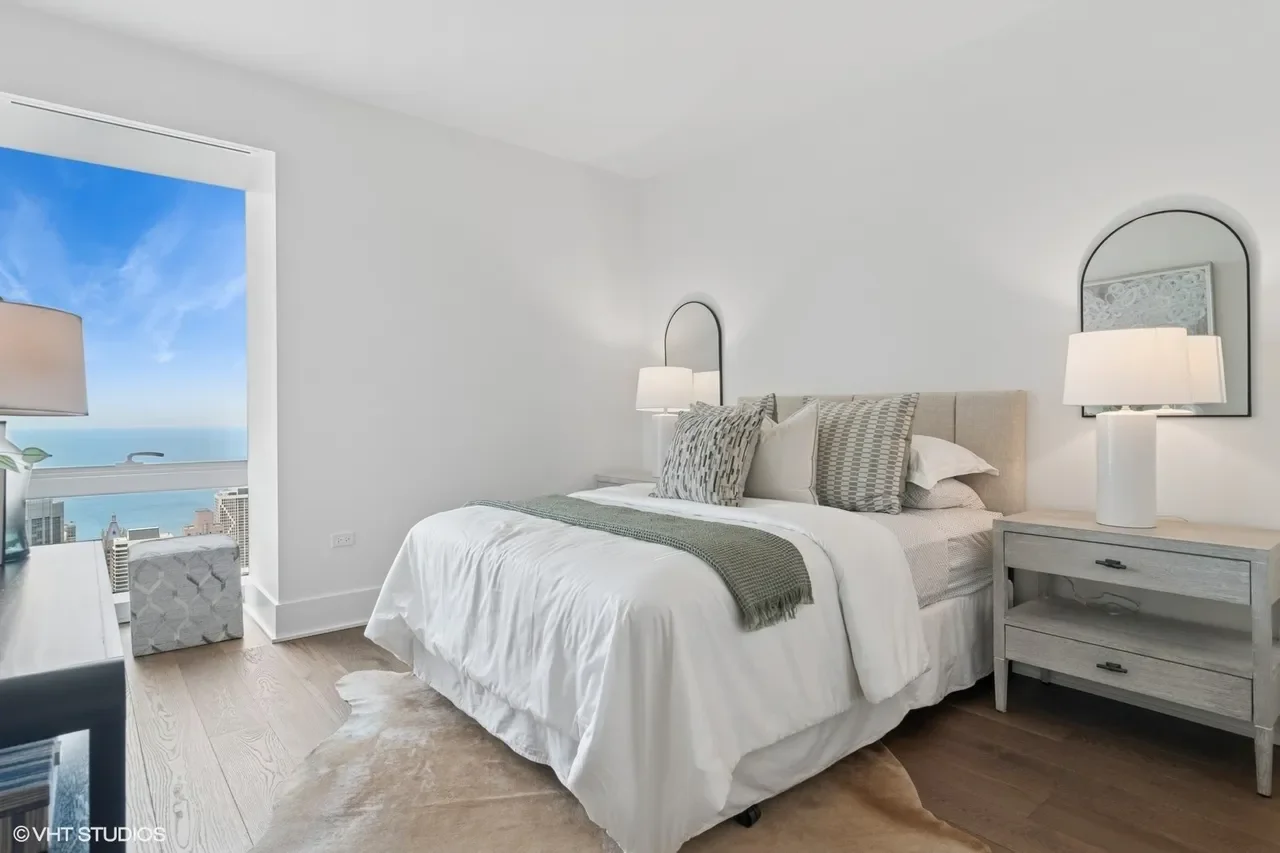 Bedroom with large bed, white bedding, and gray pillows, two nightstands with lamps, and a window showing a cityscape and ocean view.
