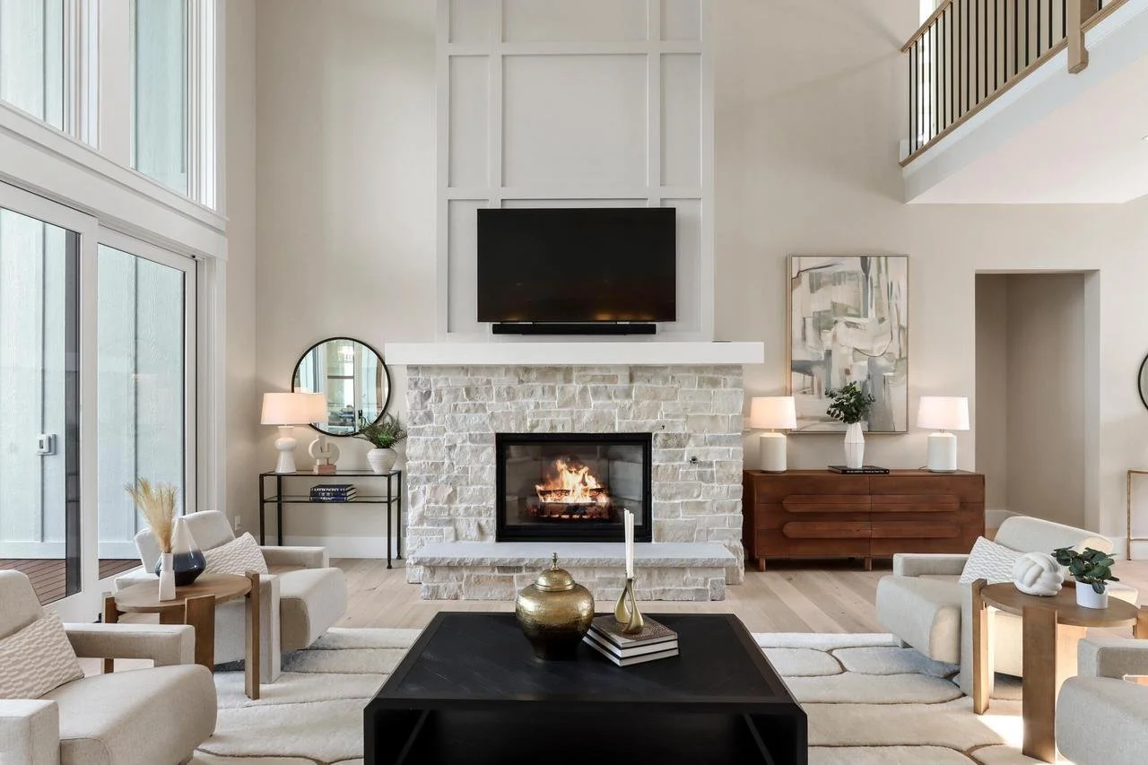 Living room with a white brick fireplace, a mounted TV above, neutral-toned furniture, side tables with lamps, and decor accents, with large windows and a high ceiling.
