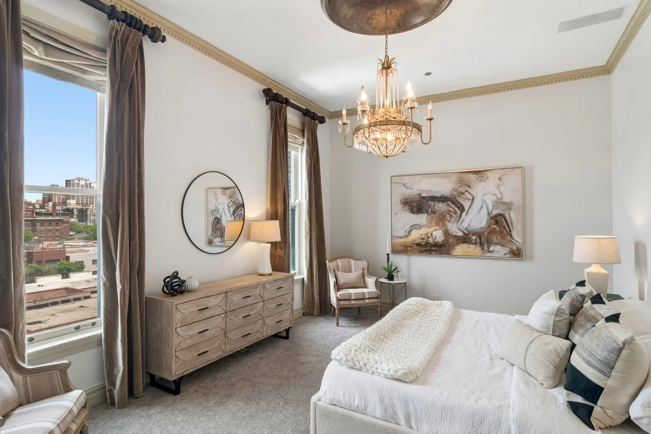 A bedroom with a bed, a chandelier, a large window with curtains, a dresser with a mirror, a lamp, a chair, and abstract wall art.