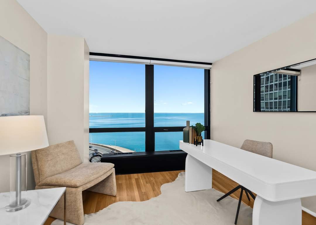 Modern hotel room with a large window view of the ocean, beige walls, a beige armchair, a white lamp, a white desk with decorative items, and a mirror on the wall.
