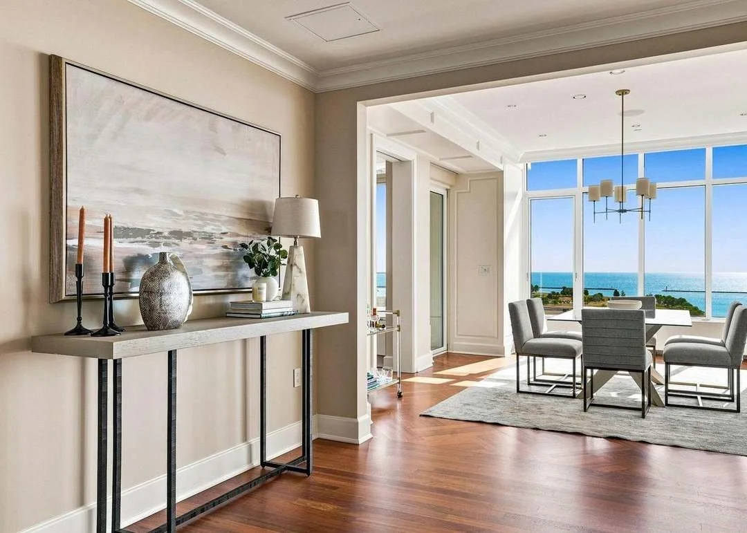 Modern dining room with large windows showing ocean view, marble table with six chairs, white walls, Hardwood floors, chandelier, and minimalist decor.
