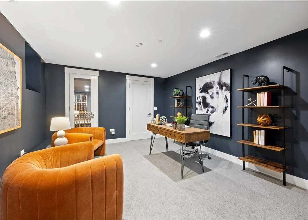 Modern office with dark blue walls, gray carpet, orange velvet seating, wooden desk, black office chair, white door with window, wall art, and wooden shelves with decorative items.