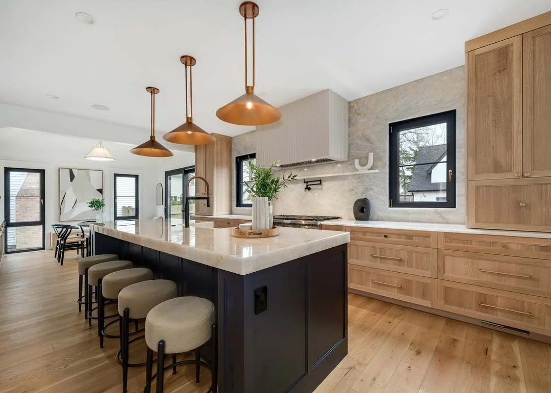 Modern kitchen with black island and three copper pendant lights above, light wood cabinetry, marble countertops, two windows, and dining area in the background.