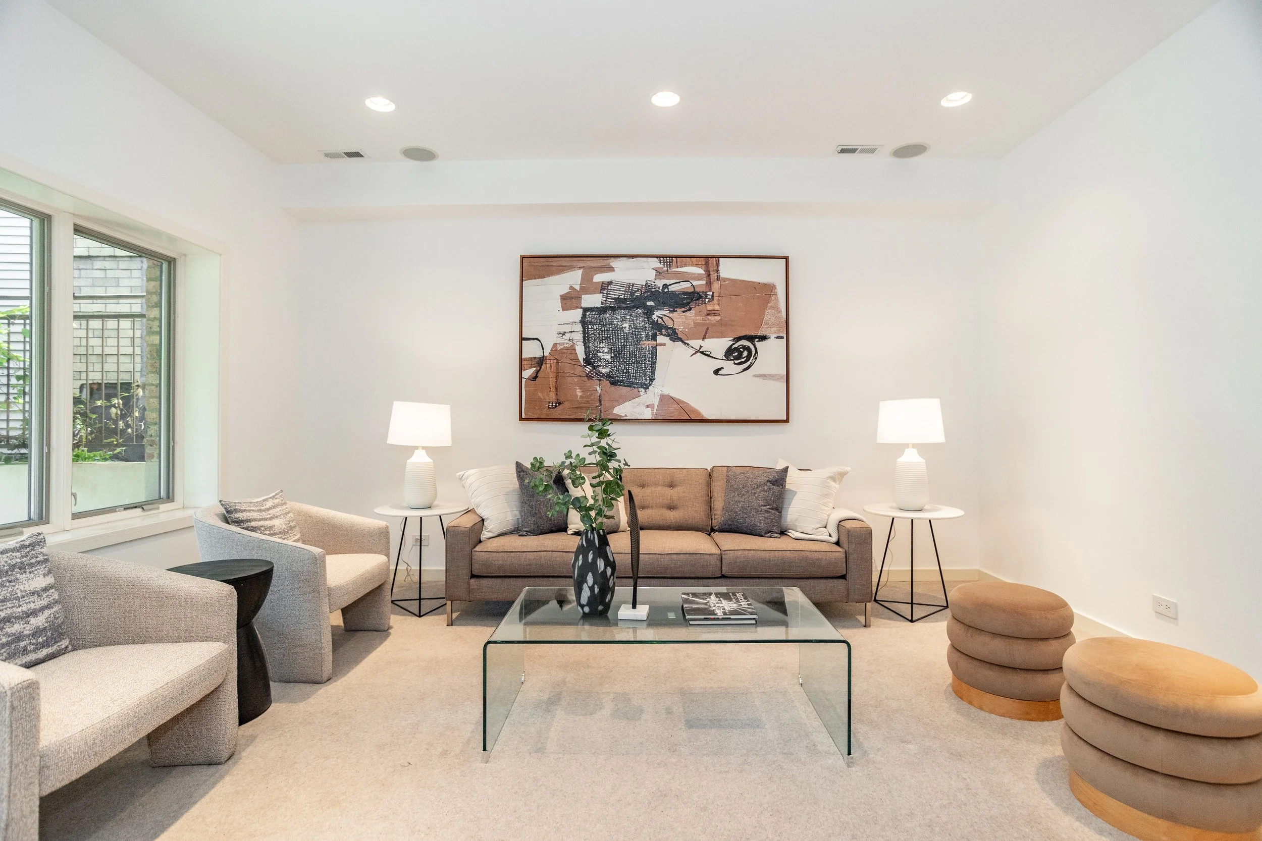 Living room with beige sofa, gray armchairs, glass coffee table, two white lamps on black side tables, abstract artwork on white wall, window with greenery outside, beige carpet, and round beige ottomans.