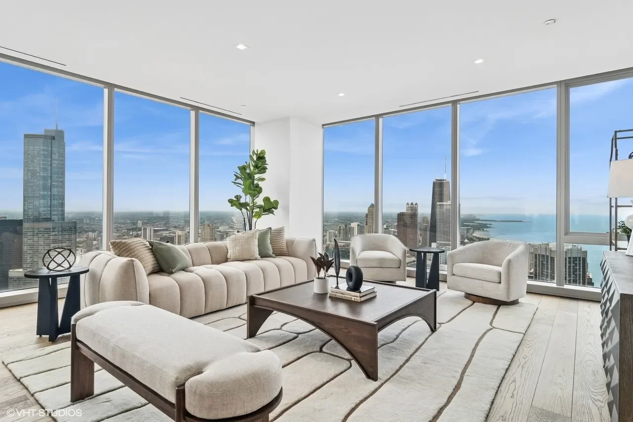 Modern living room with large floor-to-ceiling windows showing city skyline and water, beige sectional sofa, two armchairs, a wooden coffee table with decorative items, side tables, a rug, and indoor plants.