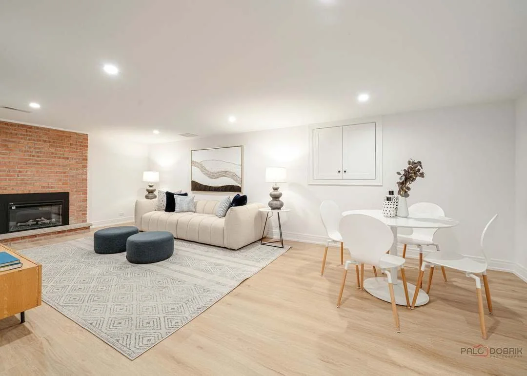 Modern living room with beige sofa, two dark ottomans, a rug, a brick fireplace, and a dining area with a white round table and six white chairs. The room is decorated with lamps, wall art, and vases on the table.