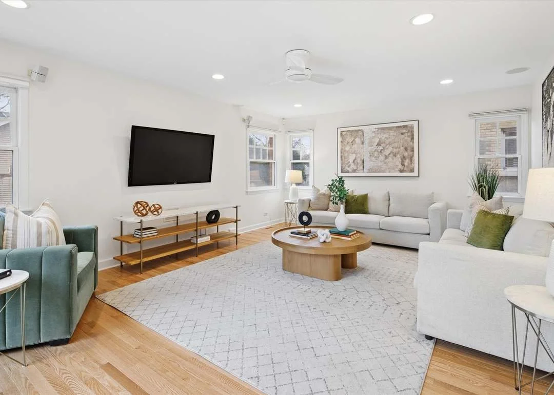 Bright living room with white walls, hardwood floors, a large white sofa with green pillows, a white armchair, a round wooden coffee table with decorative items, a minimalist TV stand with decorations, and several windows with white blinds allowing natural light.