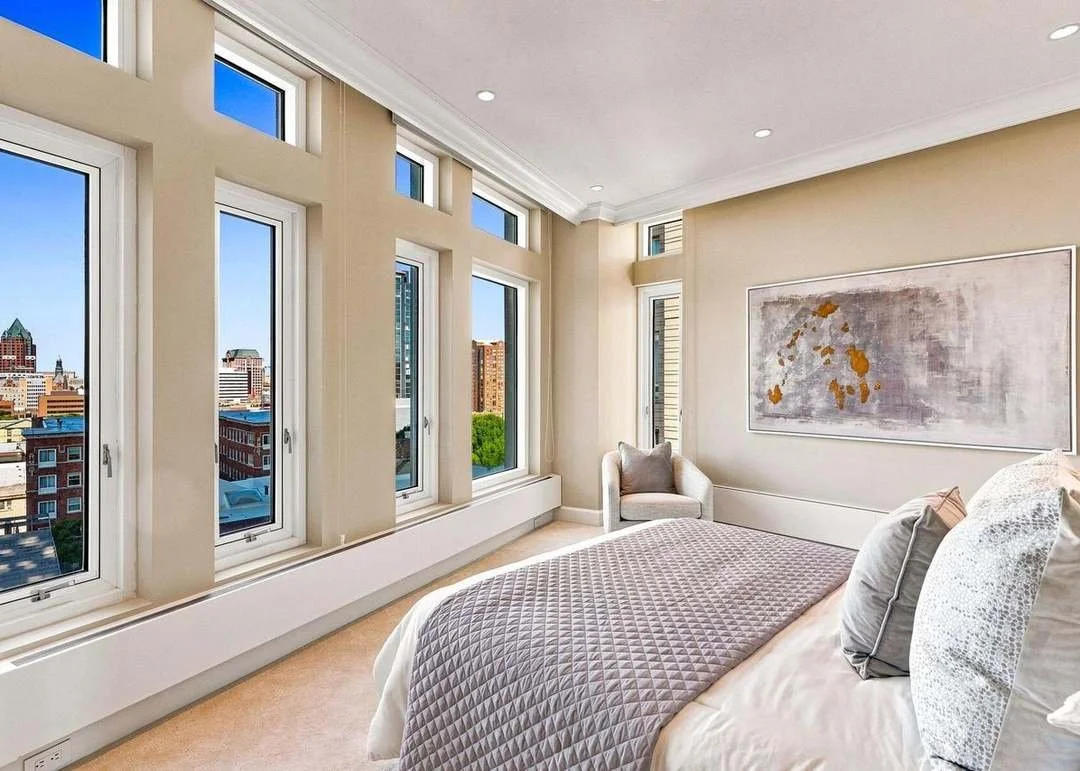 Bright bedroom with large windows showing city skyline, a bed with gray quilt, decorative pillows, a beige armchair, and abstract wall art.