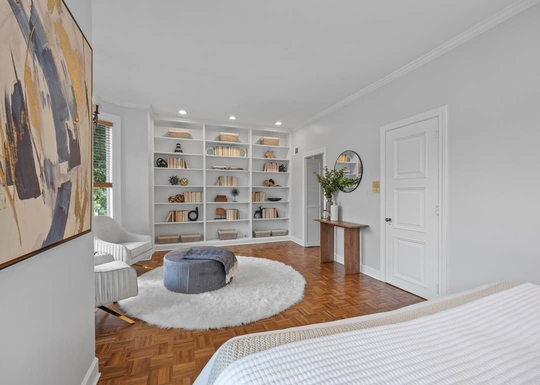 A bedroom with white walls, a white bookshelf filled with books and decorative items, a white shag rug, a light gray ottoman, a white armchair, a wooden table with a vase of flowers, a mirror, and a closed white door.