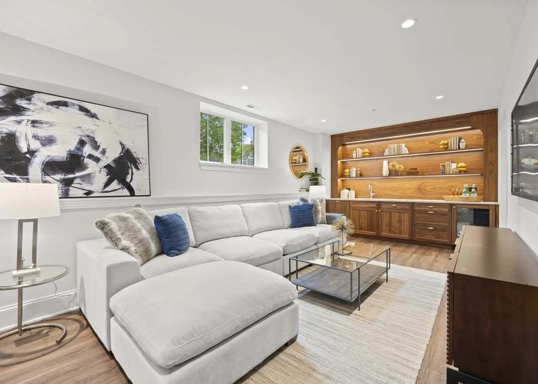 Modern living room with white sectional sofa, blue and faux fur pillows, glass coffee table, and wooden built-in shelves with books and decorative items, bright window, and abstract artwork.