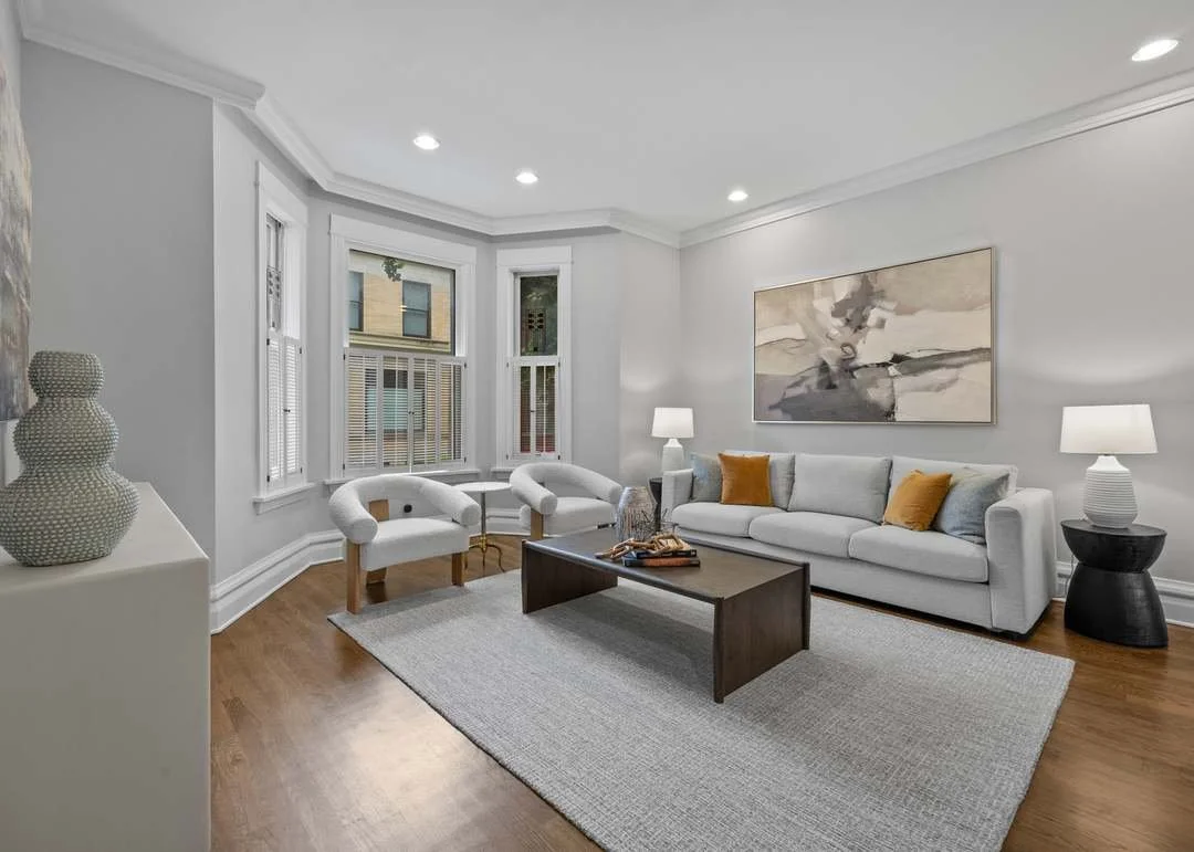 Living room with white walls, hardwood floors, a white sofa with yellow and gray pillows, a coffee table, two white armchairs near windows, and two side tables with lamps.