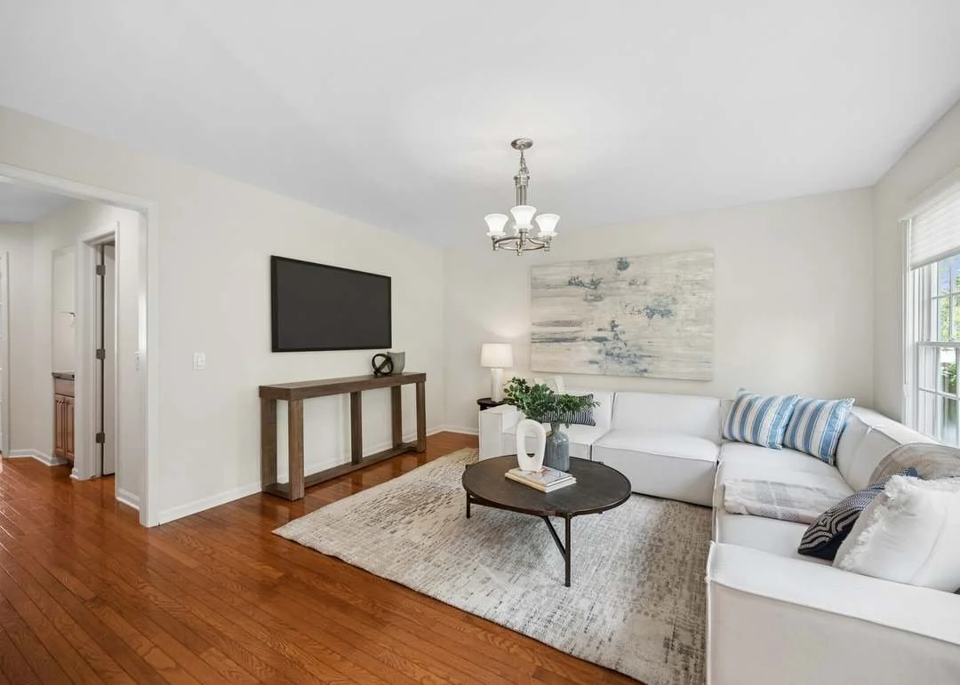 Living room with white walls, hardwood floor, a white L-shaped sofa with striped pillows, a round dark coffee table, a beige area rug, a large abstract painting, a tall white lamp, a wooden console table, a wall-mounted flat-screen TV, and a chandelier, with windows on the right side.