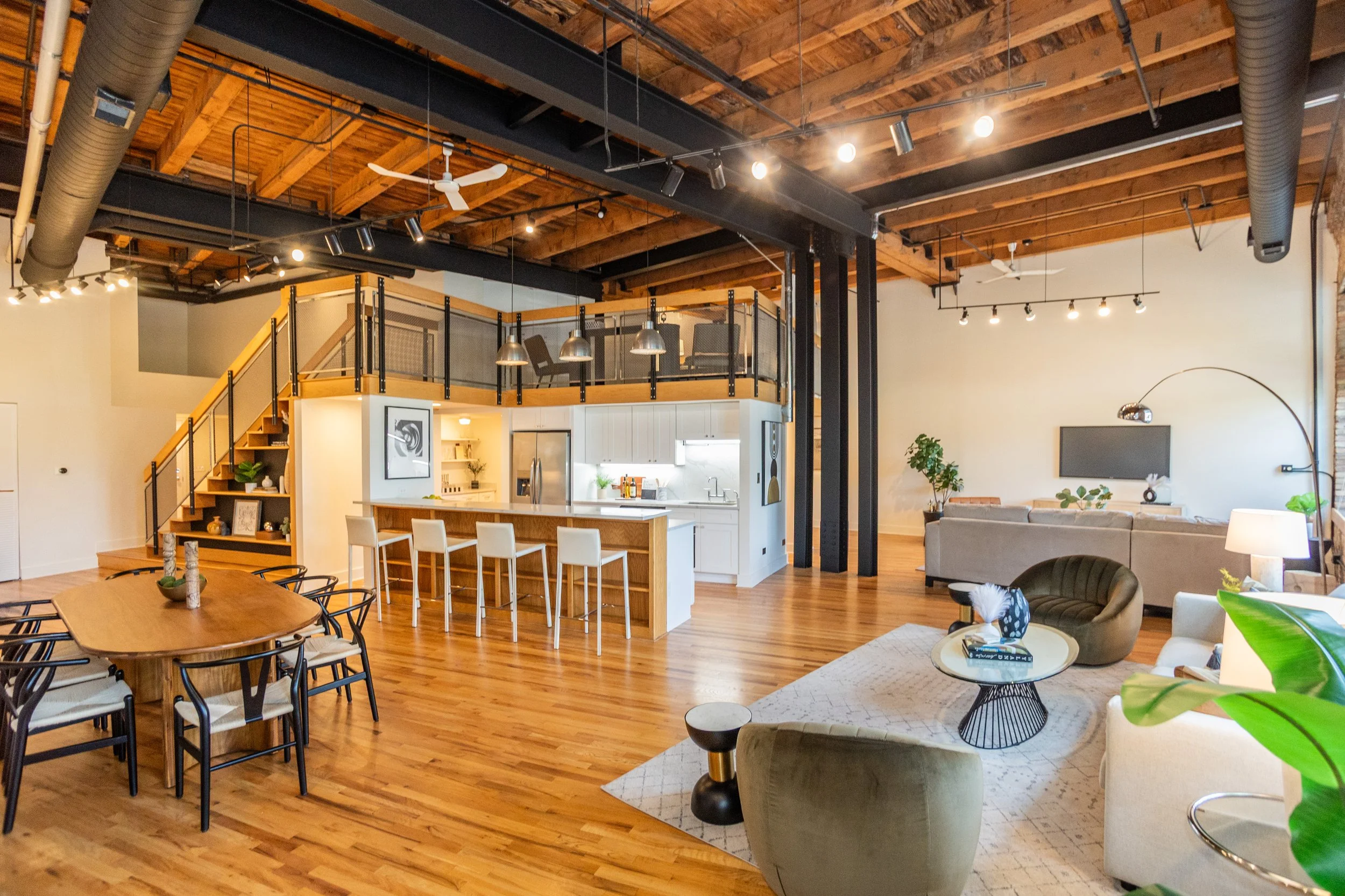 Spacious open-plan living area with a dining table, bar stools at a kitchen island, and a sitting area with sofas and chairs, featuring wooden floors, exposed wooden ceiling, and modern decor.