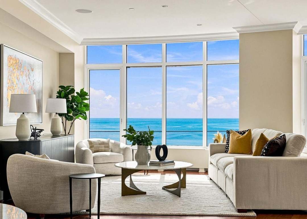 Living room with large window overlooking the ocean, white sofas with yellow and black pillows, a coffee table with a vase and books, and decorative lamps and artwork.