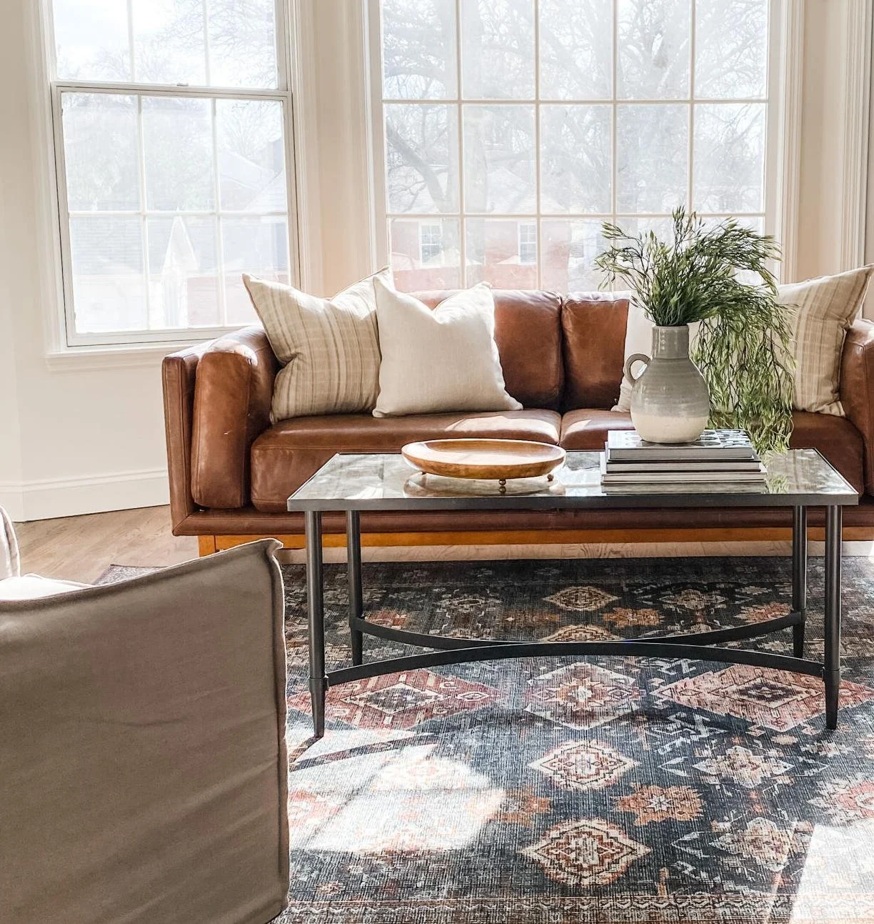 Living room staged by Olive + Opal in St. Louis with brown leather sofa, decorative pillows, glass coffee table with books and vase, patterned area rug, and large windows.