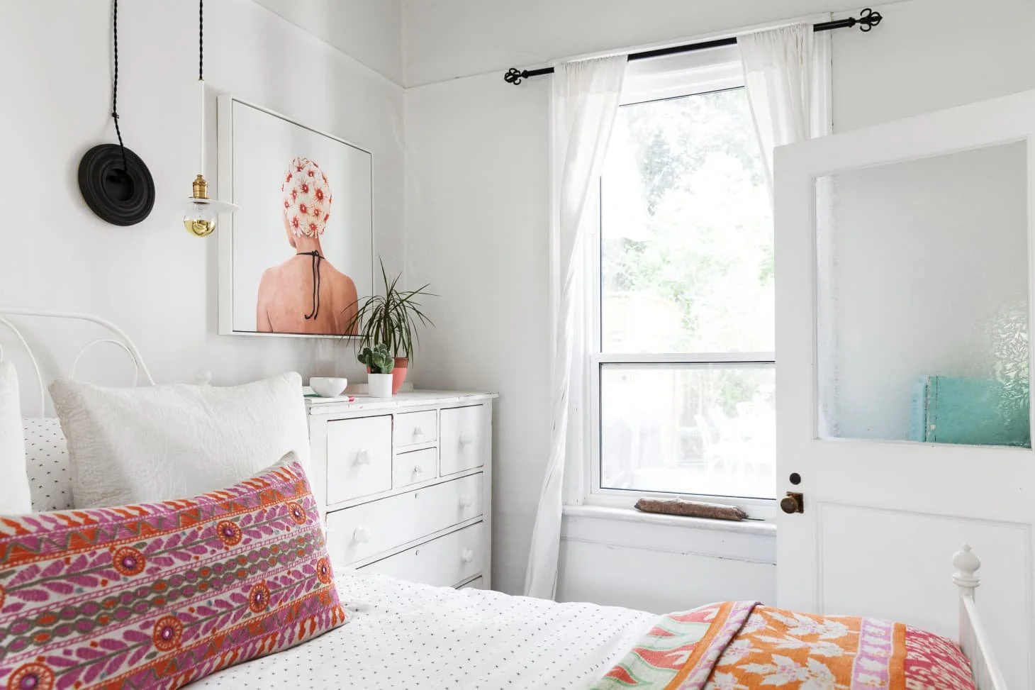 Minimalist bedroom staged in St. Louis with white decor, featuring a bed with colorful pillows, a white dresser, a plant, and a large window. A unique light fixture hangs above a framed art piece on the wall.