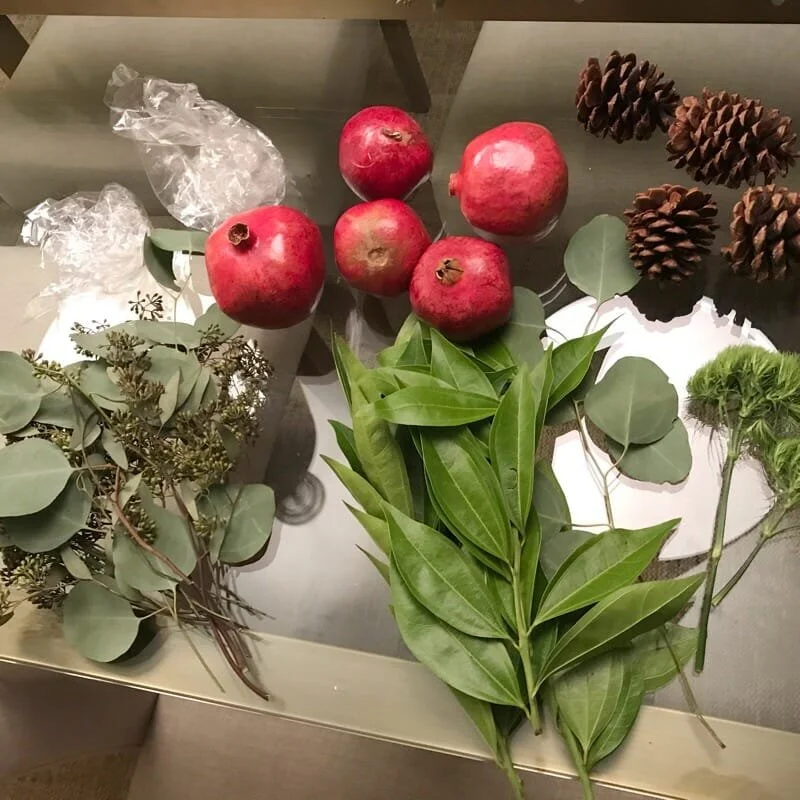 Glass table with pomegranates, eucalyptus leaves, green branches, pinecones, and clear plastic wrap used as home staging accessories.