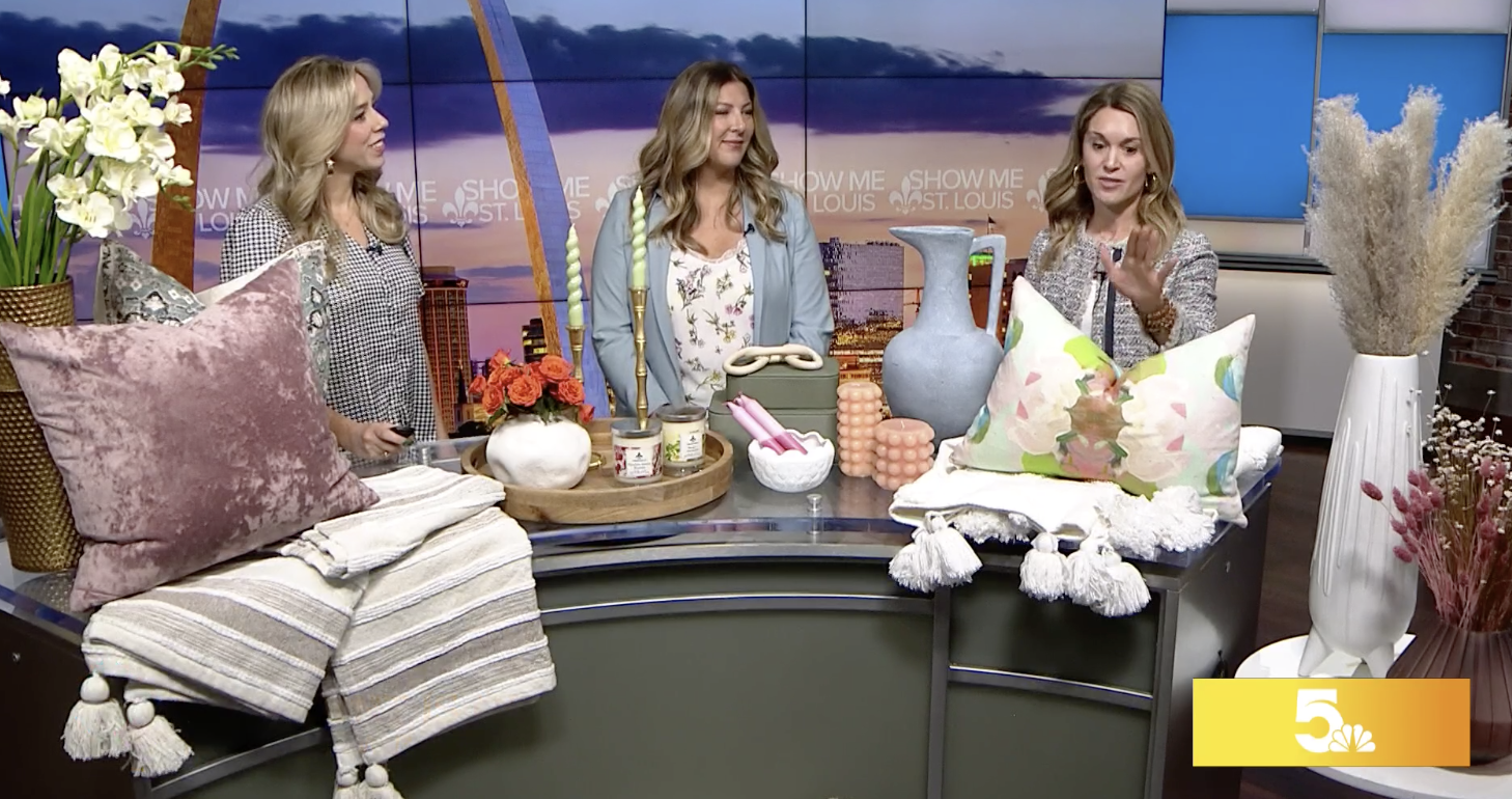 Three women stand behind a table with home decor items on a TV show set. The table displays pillows, vases, flowers, candles, and decorative items. The background includes a screen with a logo and city skyline graphics.