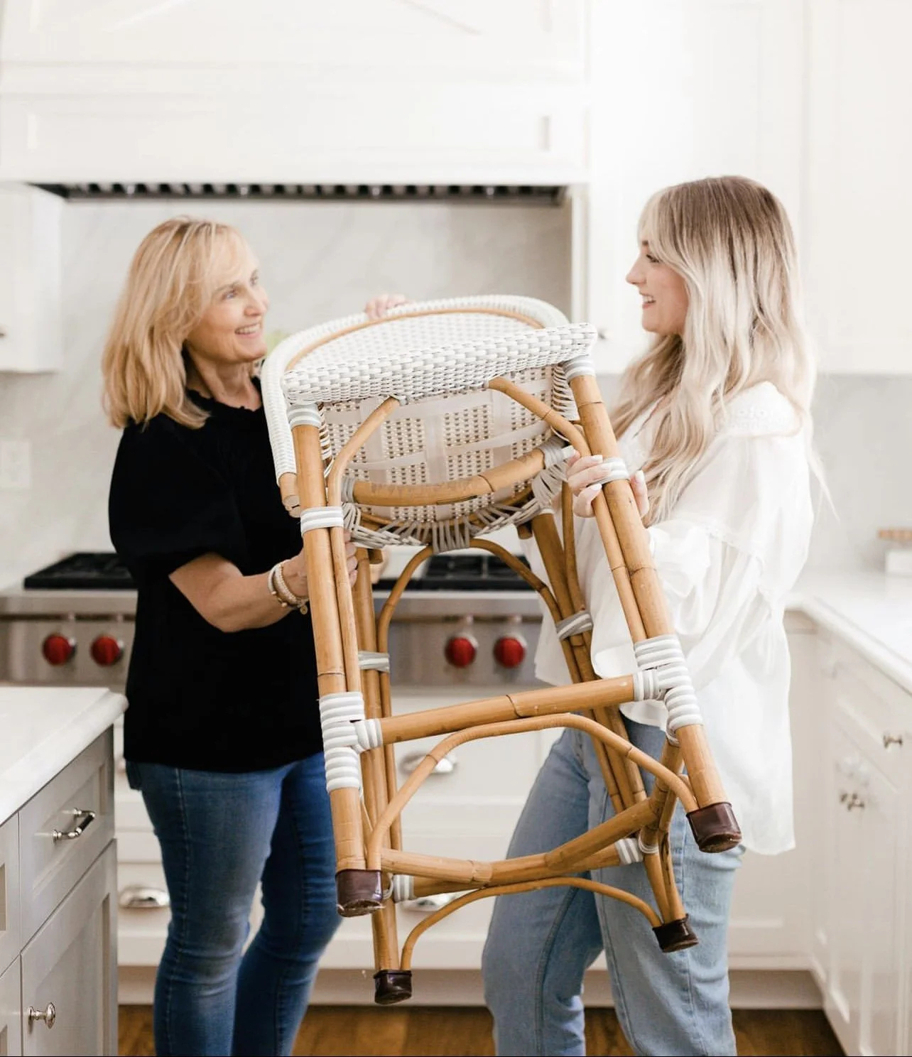 Two home staging owners smiling while lifting a wicker chair in a kitchen.