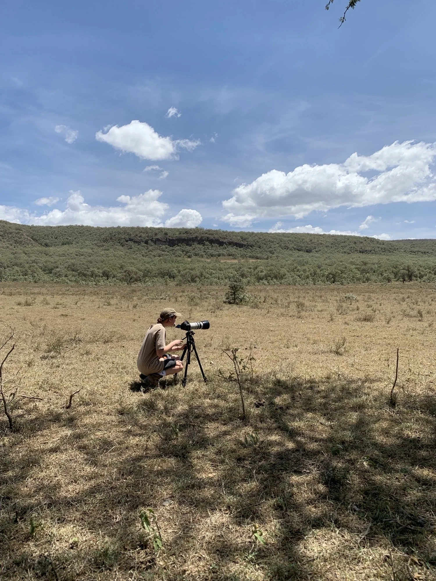 Person with a telescope sits on the ground in a dry grassy field with hills and trees in the distance under a partly cloudy sky.