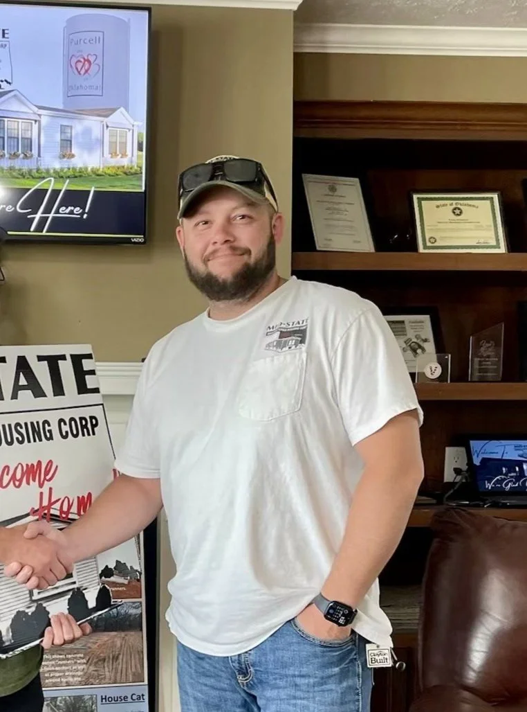 A man with a beard and mustache, wearing a white t-shirt, sunglasses on his cap, and a smartwatch, standing inside a room with a bookshelf that has framed certificates, shaking hands with someone. There is a sign, a monitor, and a framed certificate behind him.