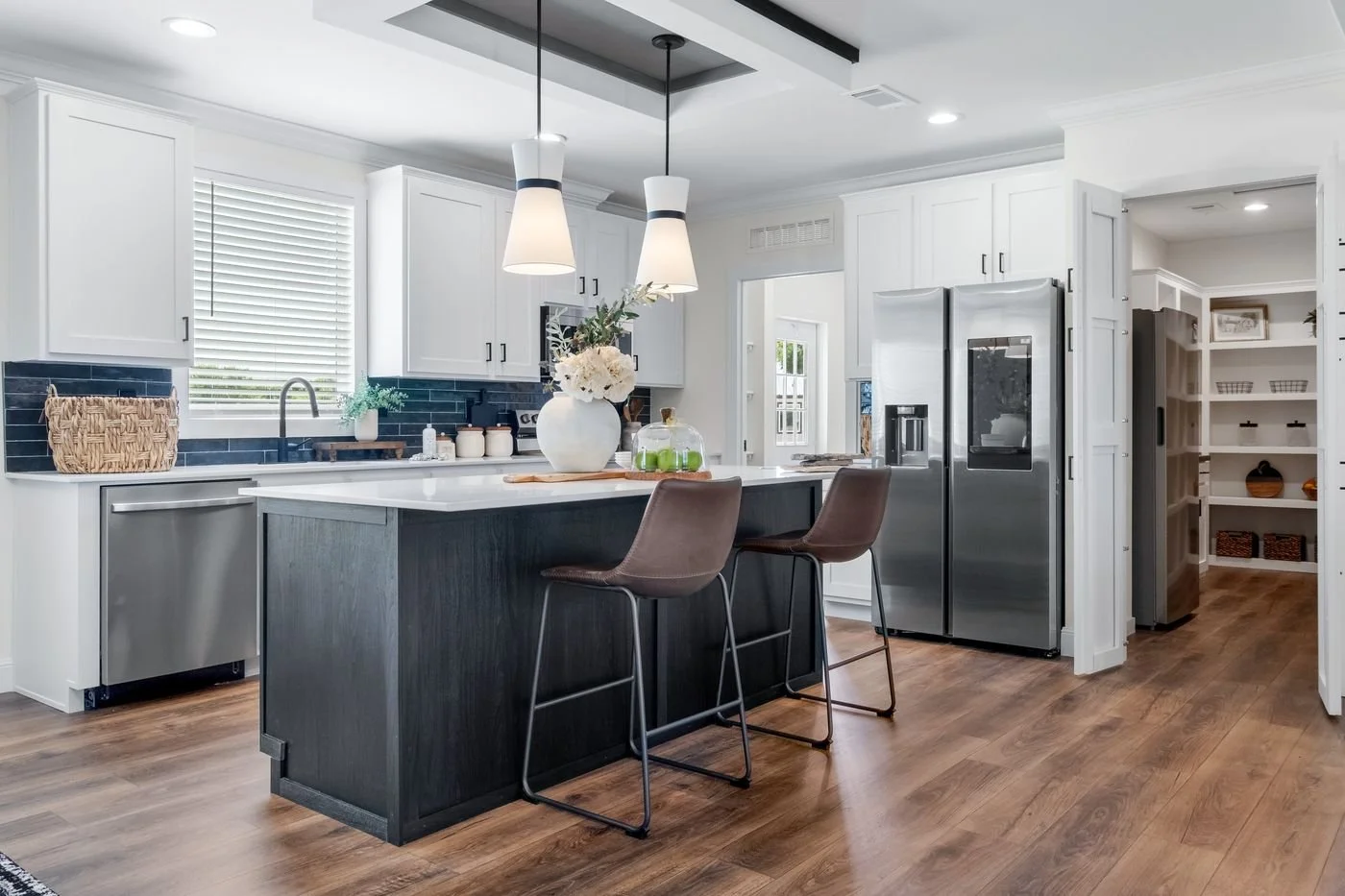 Modern kitchen with white cabinets, dark blue backsplash, stainless steel appliances, and wooden flooring. There is a kitchen island with two brown chairs, a vase with flowers, and hanging light fixtures.