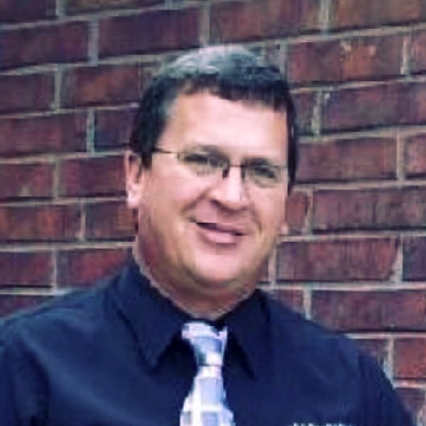 A smiling man with dark hair, glasses, wearing a black shirt and light-colored tie, standing in front of a brick wall.