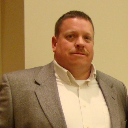 A man in a gray blazer and white shirt standing indoors against a beige wall.
