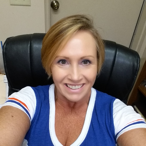 A smiling woman with short blonde hair wearing a blue and white shirt, sitting in a black leather chair in an office with a door and some papers behind her.