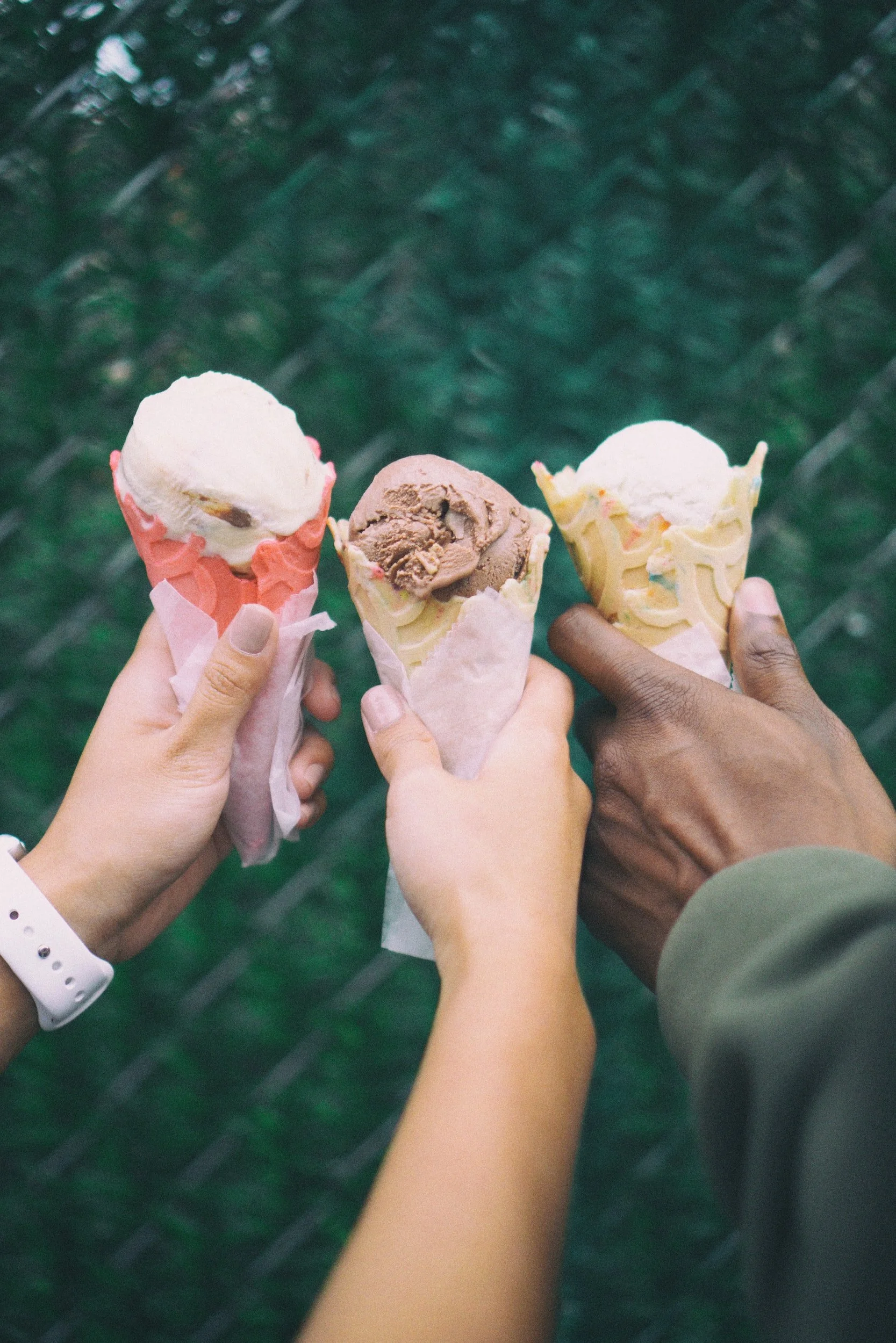Three hands holding ice cream cones with different flavors against a green background.