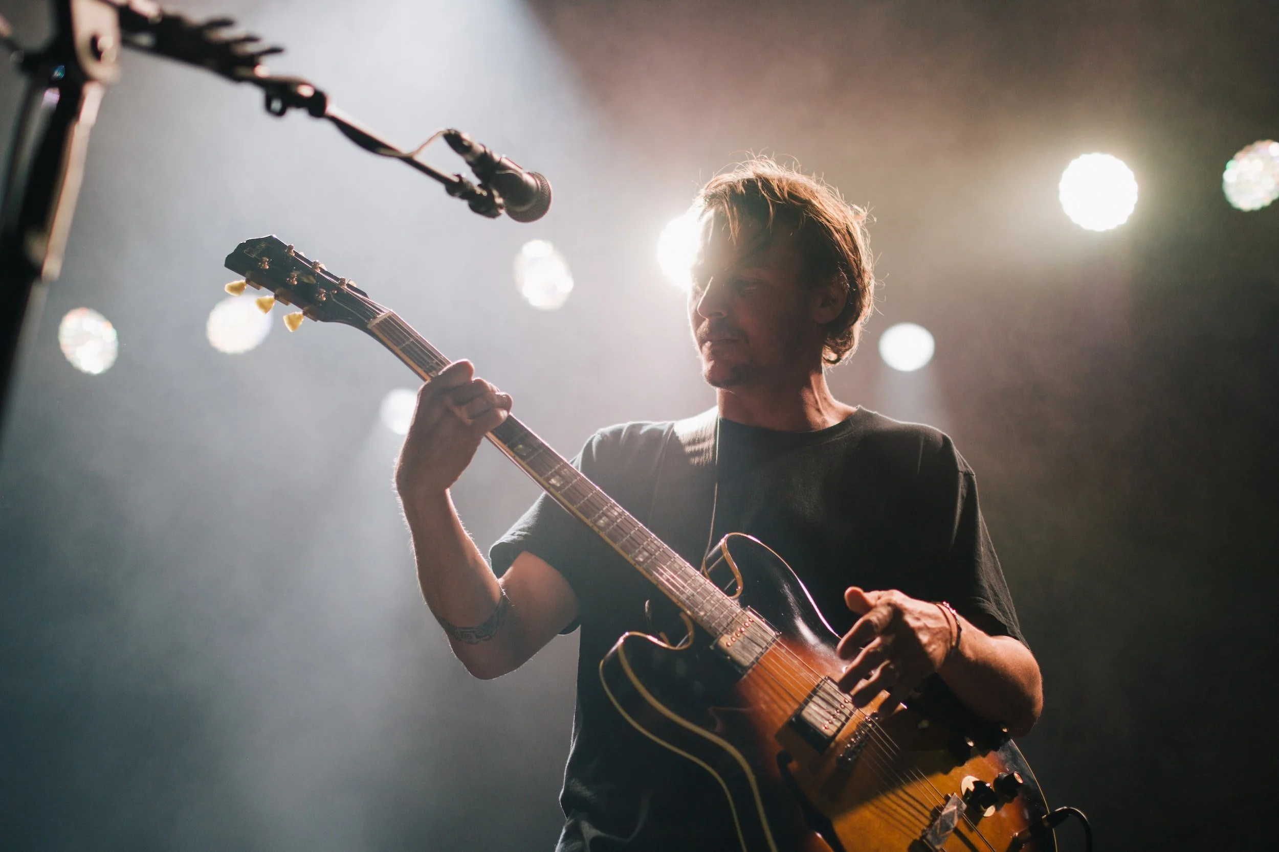 Musician playing electric guitar on stage with microphone overhead and stage lights in the background.