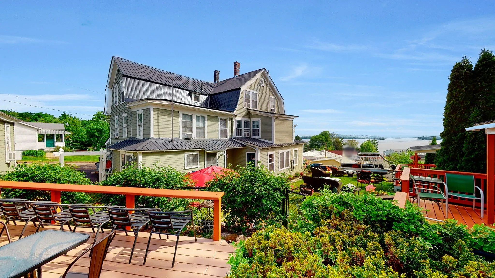 Large multi-story house with a metal roof, surrounded by greenery and a deck with chairs, under a clear blue sky.
