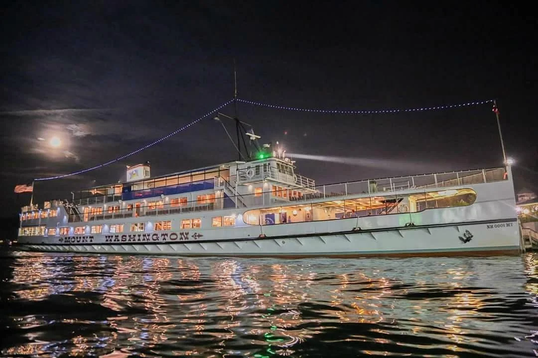 A large passenger boat named Mount Washington, lit up at night with string lights, cruising on the water under a bright moon.