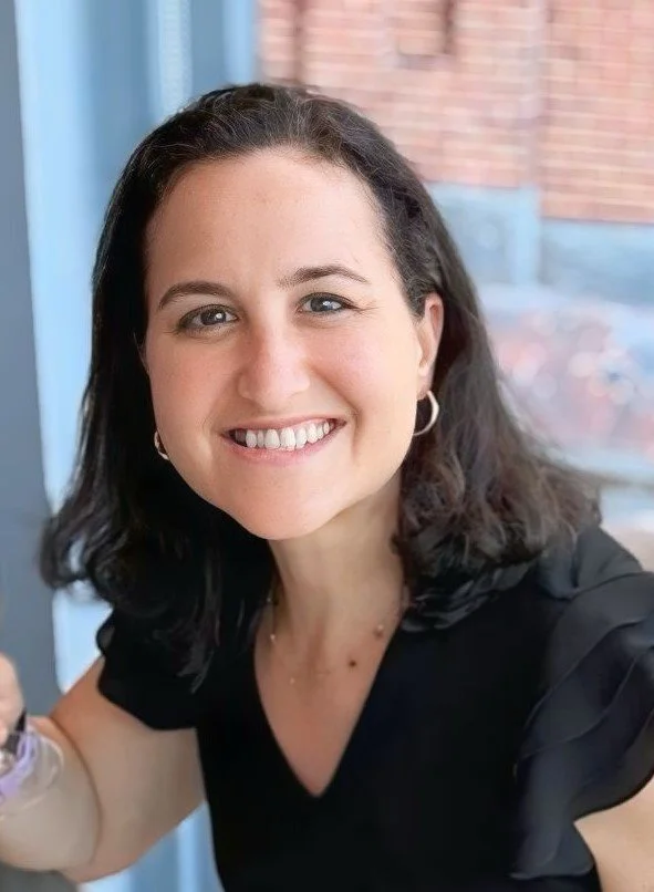 A woman with dark brown hair, smiling, wearing hoop earrings and a black top, sitting near a window with a brick building in the background.