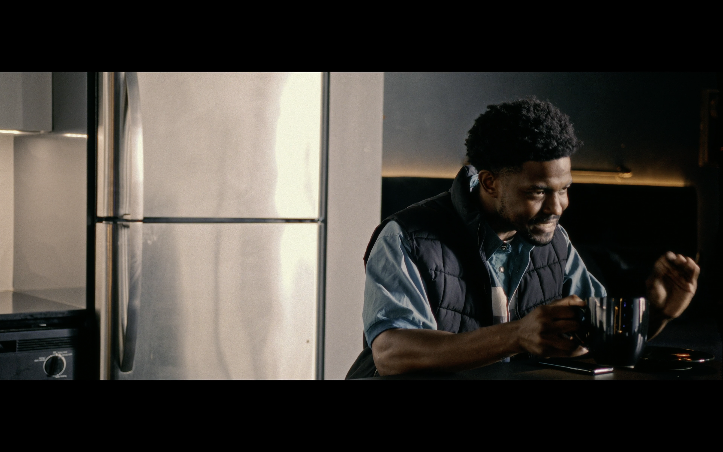 A man with curly black hair, wearing a blue shirt and black vest, sitting at a kitchen table, holding a mug.