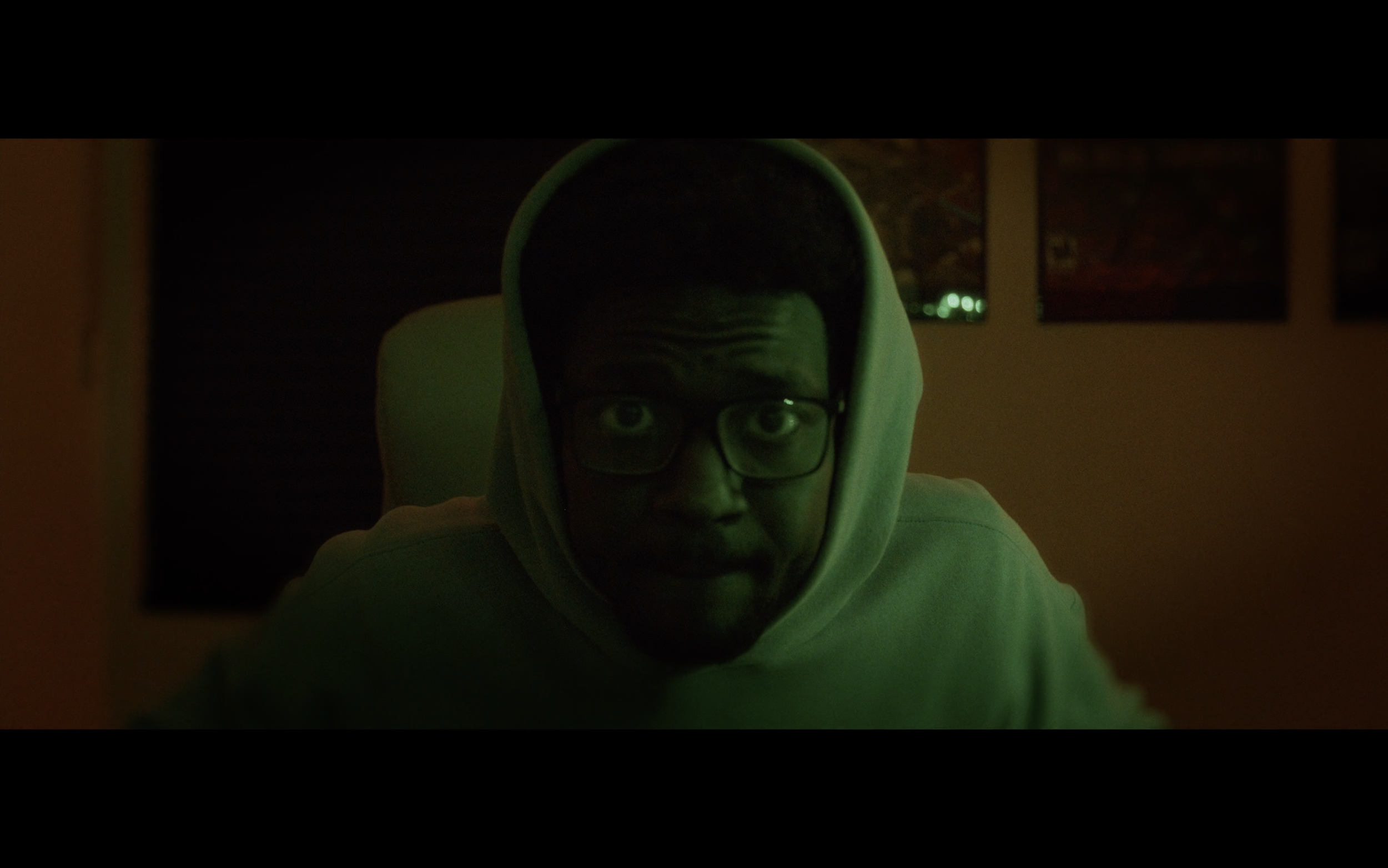 A person wearing glasses and a hooded sweatshirt, looking directly at the camera with a serious expression in a dimly lit room.