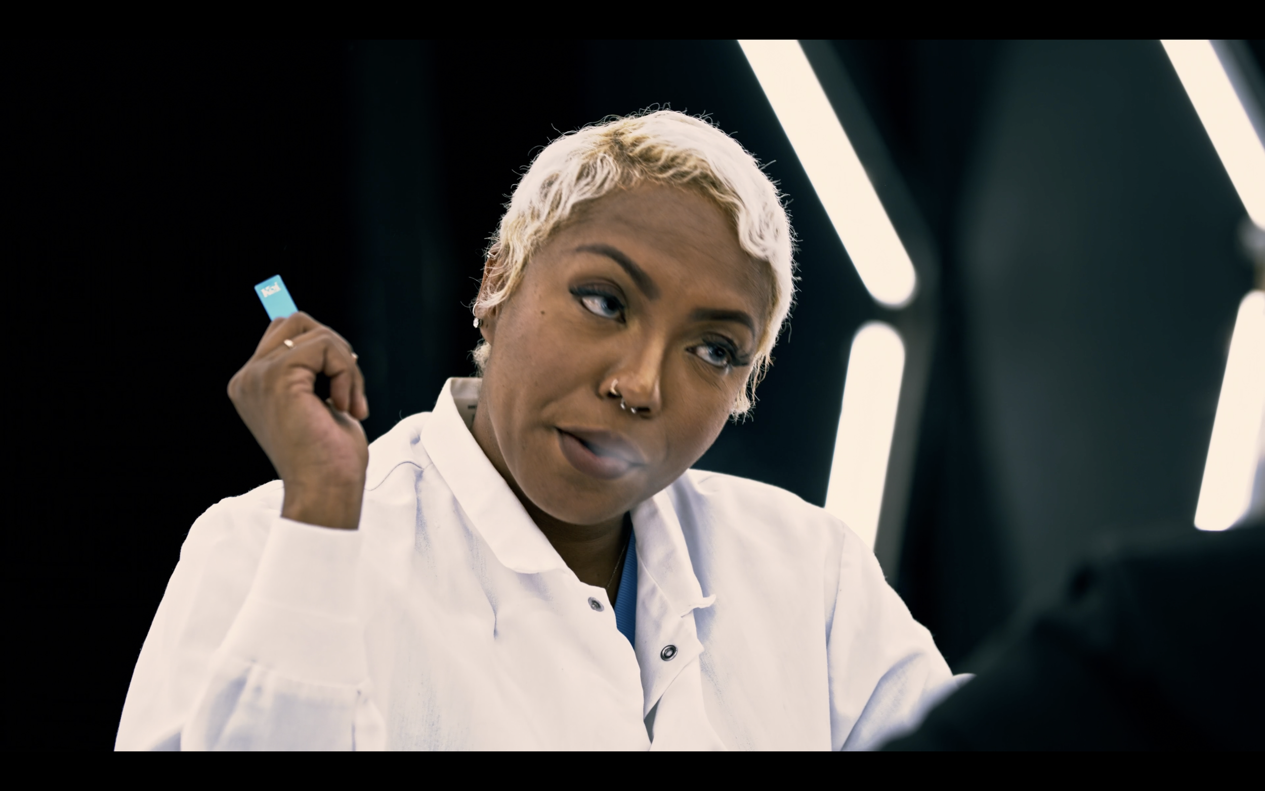 A woman with short blonde hair and multiple nose piercings, wearing a white lab coat, holding a credit card in her right hand, with a focused expression, in a modern setting with bright vertical lights.