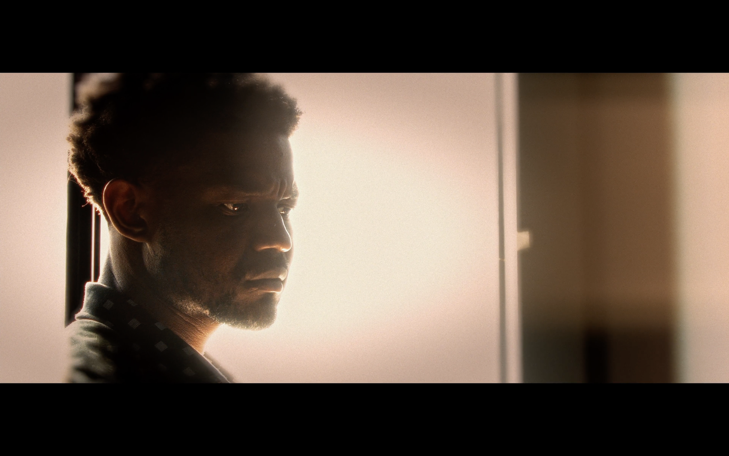 A young man with curly hair and a serious expression looking towards the right, illuminated by soft light from a window.