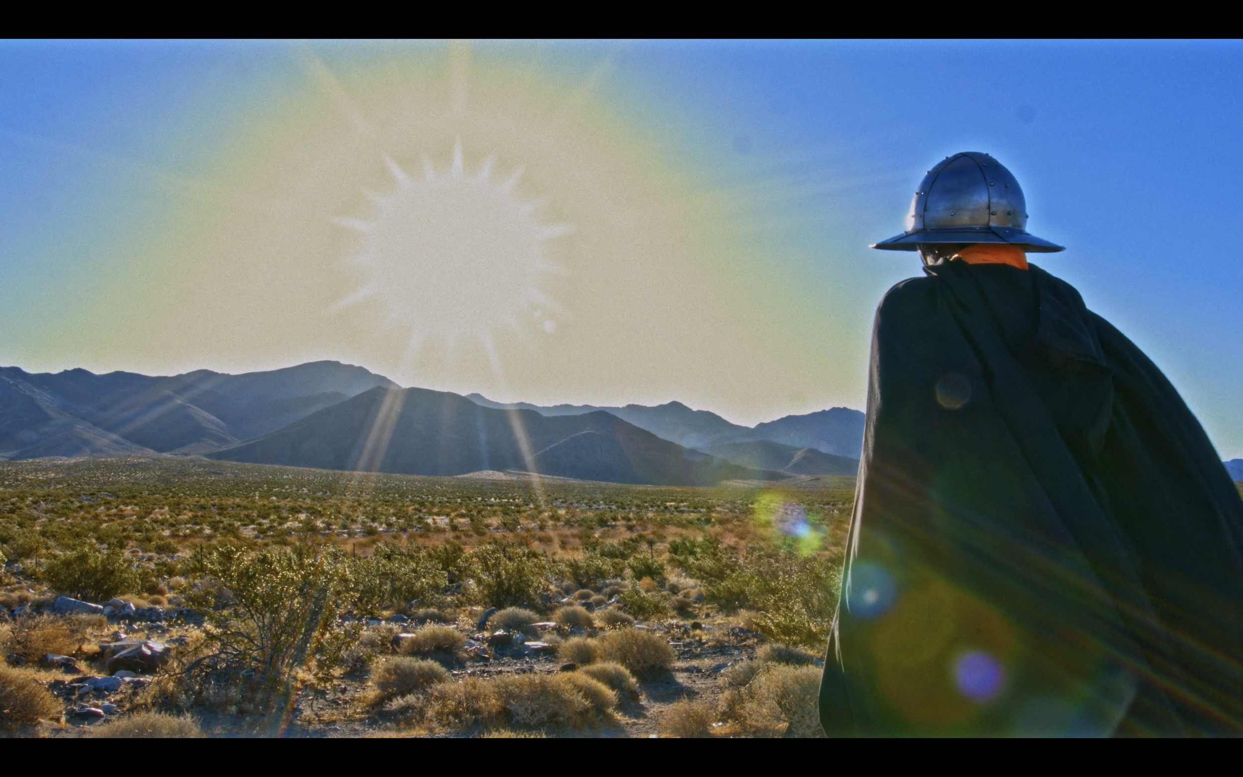 A person wearing a metal helmet standing in a desert landscape with mountains in the background, under a bright sun.
