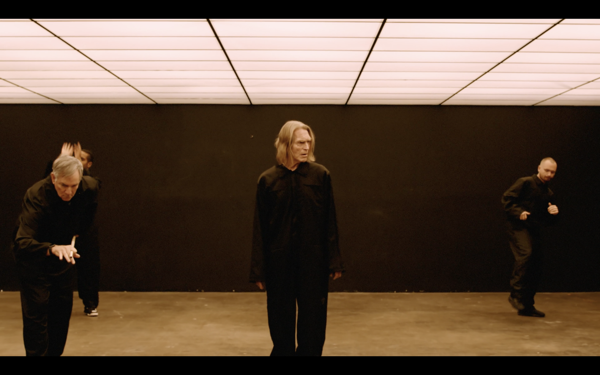 Four people dressed in black participating in a dance or performance in a dimly lit room with a dark wall and ceiling lights.