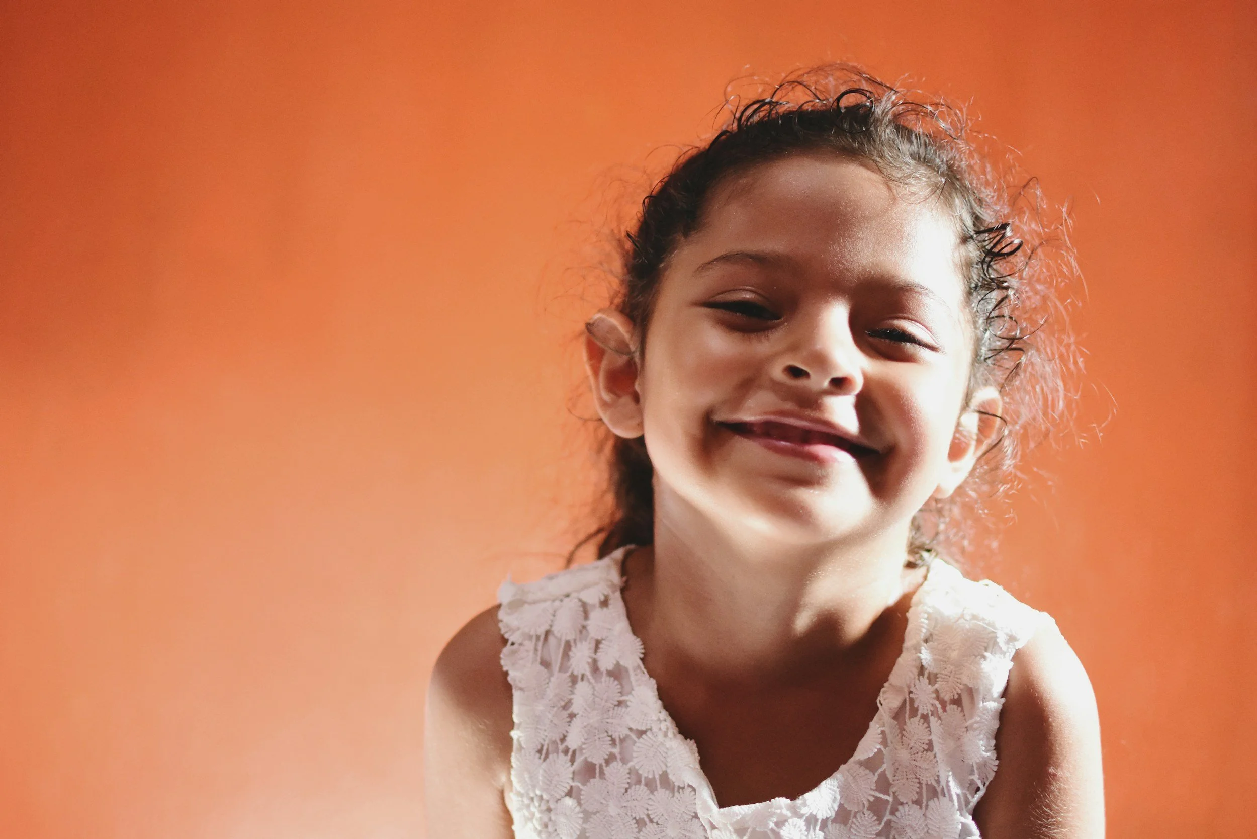 A young girl with curly dark hair smiling in front of an orange background, wearing a white lace top.